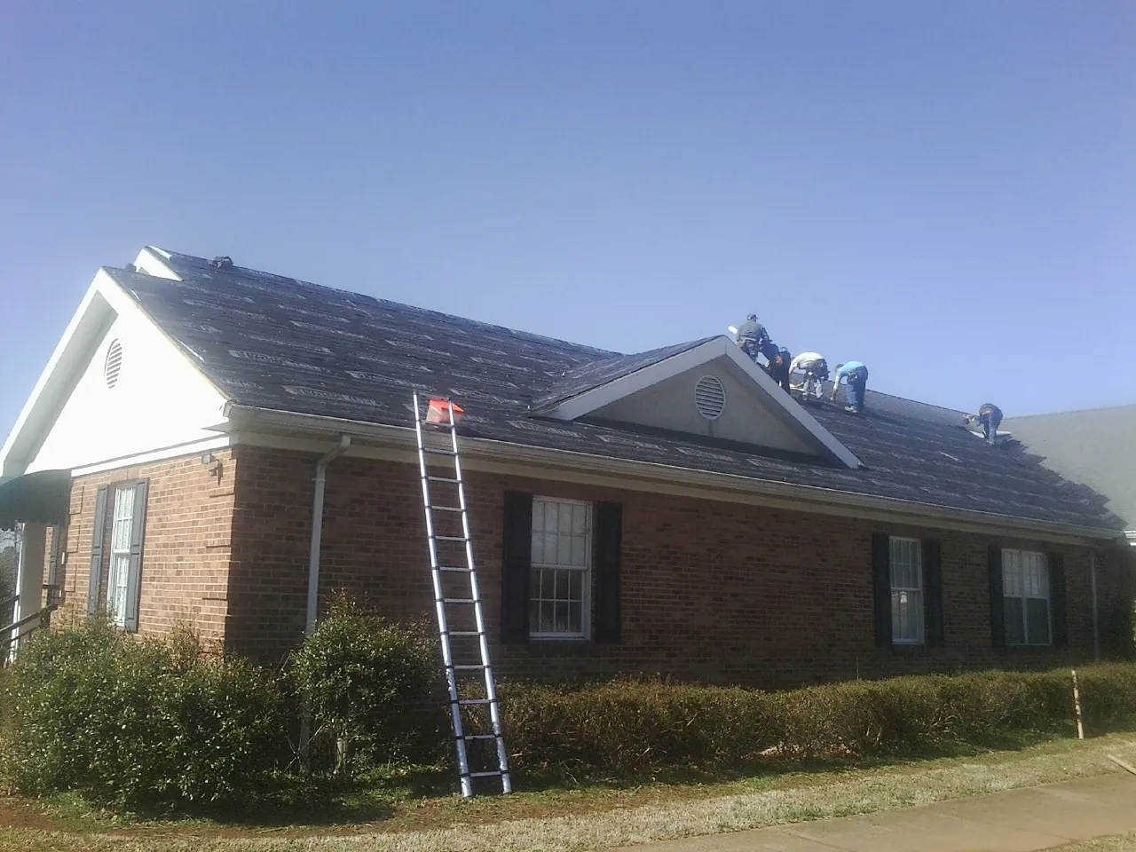 Crew tearing shingles off a brick ranch roof with a ladder set — Emergency Roof Repair work by our crew in Sudbury
