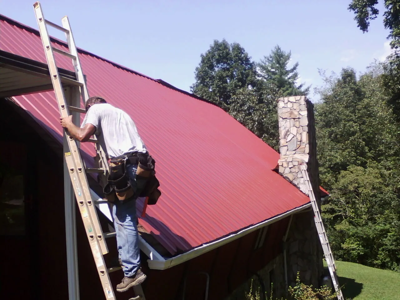 Roofer on a ladder beside a red metal roof and stone chimney — Soffit & Fascia Repair by Roofing Brothers in Sudbury